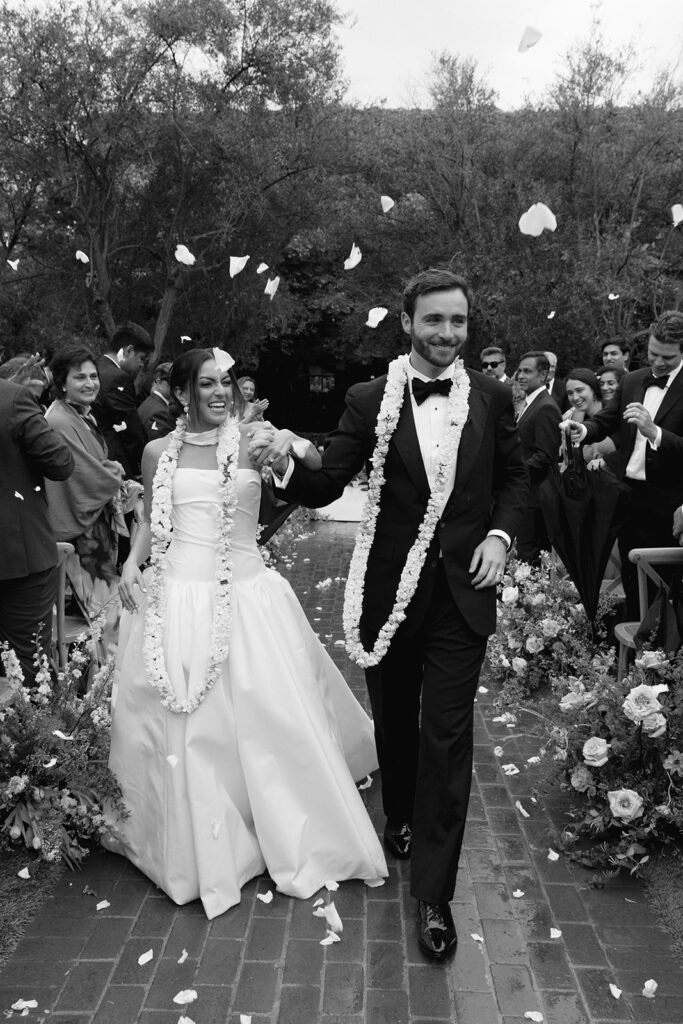 Floral garlands brushed their shoulders as petals scattered across the brick path, a soft blur of motion behind them. The crowd rose as they walked back up the aisle, petals catching in her dress and hair.